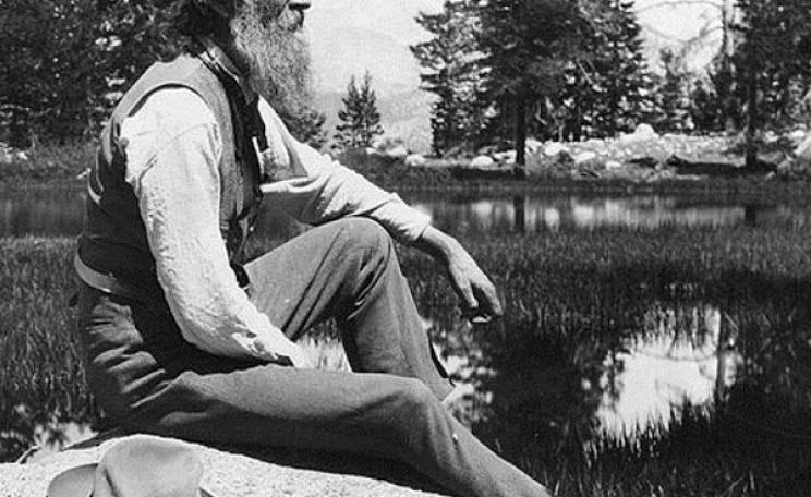 John Muir, full-length portrait, facing right, seated on rock with lake and trees in background, circa 1902. Photo: unknbown via Library of Congress (Public Domain).